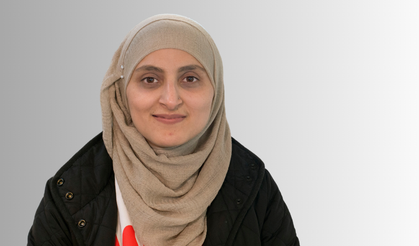 A woman smiling, she's wearing a light brown headscarf and a black jacket.